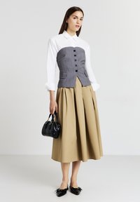 Donna che indossa una camicia bianca con corsetto a righe grigie sopra, una gonna midi beige, ballerine nere a punta e tiene una piccola borsa nera.