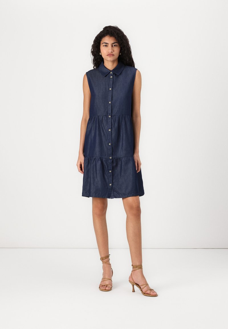Denim sleeveless dress with a collar, button-front, tiered design, and a dark blue color. Model wears gold strappy heels.