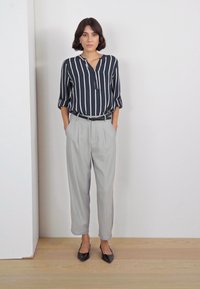 Camisa de rayas en azul marino y blanco, mangas enrolladas; pantalones grises claros, corte a medida con pliegues; bailarinas negras. Fondo neutro.