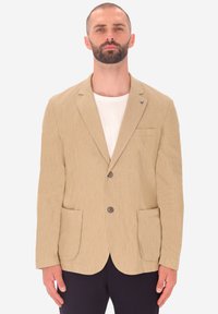 Blazer beige in tessuto con due tasche frontali, revers a punta e tre bottoni. Indossato sopra una maglietta di colore chiaro.
