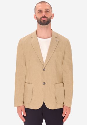 Blazer beige in tessuto con due tasche frontali, revers a punta e tre bottoni. Indossato sopra una maglietta di colore chiaro.