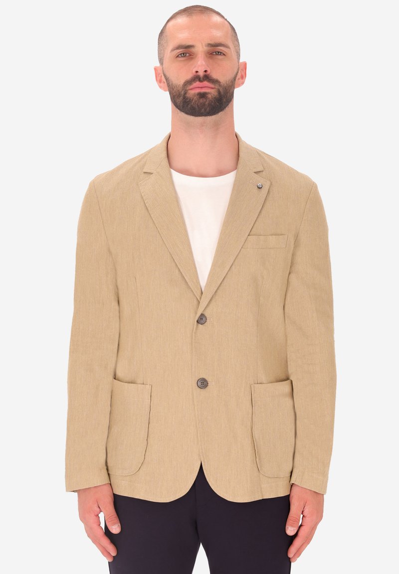 Blazer beige in tessuto con due tasche frontali, revers a punta e tre bottoni. Indossato sopra una maglietta di colore chiaro.