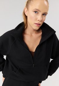 Mustast lühendatud dressipluus kõrge krae, poollukuga esiosas ja esitaskuga. Pehme kangas, iga päev kandev lõige ja minimaalsed õmblused.