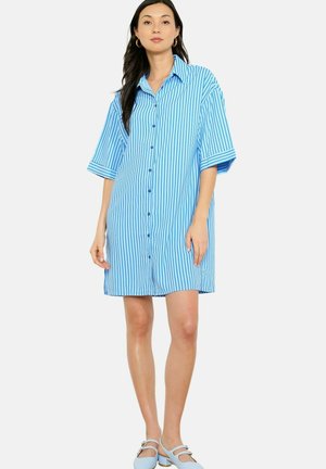 Žena s dlhými tmavými vlasmi, ktorá má na sebe modro-biele šaty na gombíky s vertikálnymi pruhmi a svetlomodré sandále s dvomi remienkami, stojaca na bielom pozadí.