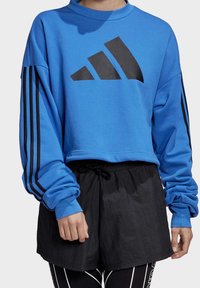 Sweat-shirt bleu court avec logo Adidas noir, col rond côtelé et manches longues avec bandes noires. Porté avec un short noir à cordon de serrage.
