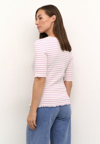 Stribet kortærmet top i hvid, lyserød og lilla. Har bølgede kanter og en tætsiddende silhuet. Parret med lyseblå jeans.