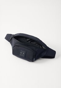 La banane en tissu navy dispose d'un compartiment principal zippé et d'une poche avant, ornée du logo "AX", avec une sangle ajustable.