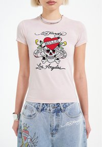 Světle růžové přiléhavé tričko s grafickým designem znázorněním lebky, zkřížených kostí a červeného srdce. Text zahrnuje "Ed Hardy", "Láska pomalu zabíjí" a "Los Angeles".