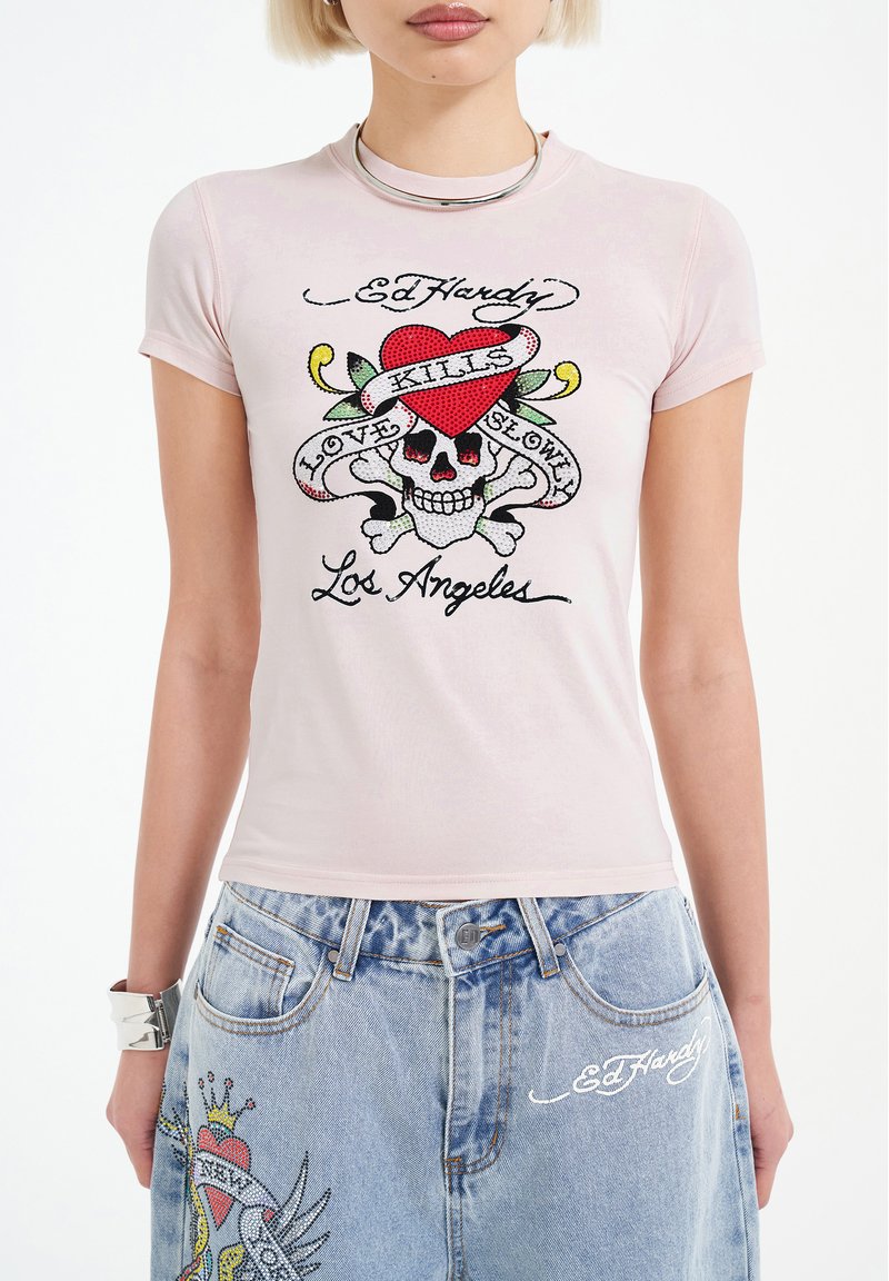 Světle růžové přiléhavé tričko s grafickým designem znázorněním lebky, zkřížených kostí a červeného srdce. Text zahrnuje "Ed Hardy", "Láska pomalu zabíjí" a "Los Angeles".