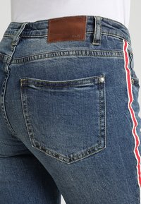Blå denimjeans med en brun läderlapp i midjan och röda och vita ränder längs sid sömmarna. Har standard bakfickor.