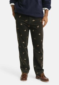 Man in donker corduroy broek met geborduurde vliegende eenden, bruine leren laarzen, marineblauwe fleece pullover en zichtbare witte mouwen van een shirt.