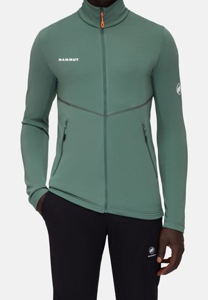 Mężczyzna w zipowanej kurtce Mammut w kolorze szałwiowym z wysokim kołnierzem oraz czarnych spodniach z logo Mammut na udzie, stoi z jedną ręką w kieszeni.