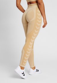 Beige Leggings mit strukturierten Mustern und hohem Bund, verziert mit seitlichen Chevron-Akzenten. Kombiniert mit weißen Sportschuhen.