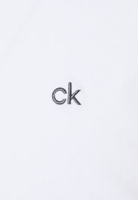 Biele tkanina s jemnou textúrou s malým vyšitým logom "ck" v šedej farbe. Jednoduchý dizajn bez ďalších vzorov alebo akcentov.