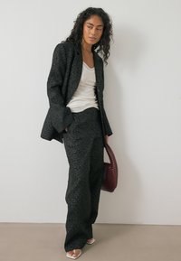 Zwart-gepatenteerde blazer en wijde broek in textiele stof, gecombineerd met een witte top en een bordeaux kleurige geweven tas. De schoenen zijn doorzichtige haksandalen.