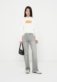 Felpa bianca con cappuccio con "TOMMY JEANS" in arancione, jeans grigi a gamba larga, scarpe bianche e una piccola borsa nera. Abbigliamento semplice e moderno.