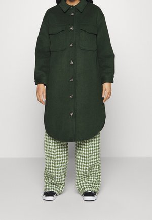 Persoon met een donkergroene, knoopgesloten lange jas over groene en witte pied-de-poule broek en zwarte sneakers, staand tegen een effen achtergrond.