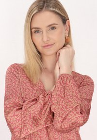 Rød bluse med blomsterprint, lange puffede ærmer og V-udskæring, lavet af let, blødt stof, båret med runde orange øreringe.