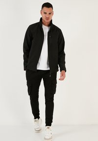 Chaqueta negra de softshell con cuello alto, cierre de cremallera y bolsillo en el pecho, combinada con pantalones cargo negros y zapatillas altas blancas.