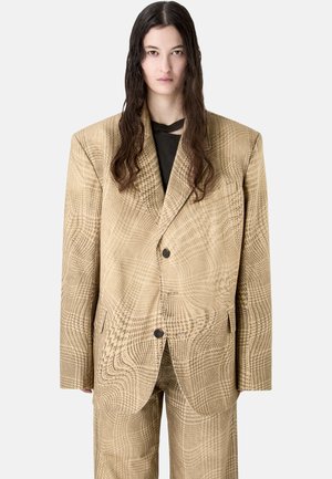Persona con capelli lunghi e scuri che indossa un tailleur oversize beige a quadri con bottoni neri e una camicia nera sotto.