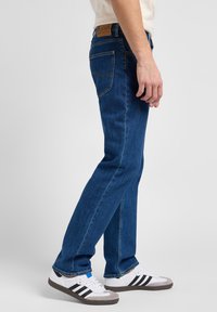 Blå denimjeans med rak passform, med bakfickor, gula sömmar och ett märkespatch i brunt läder vid midjan.