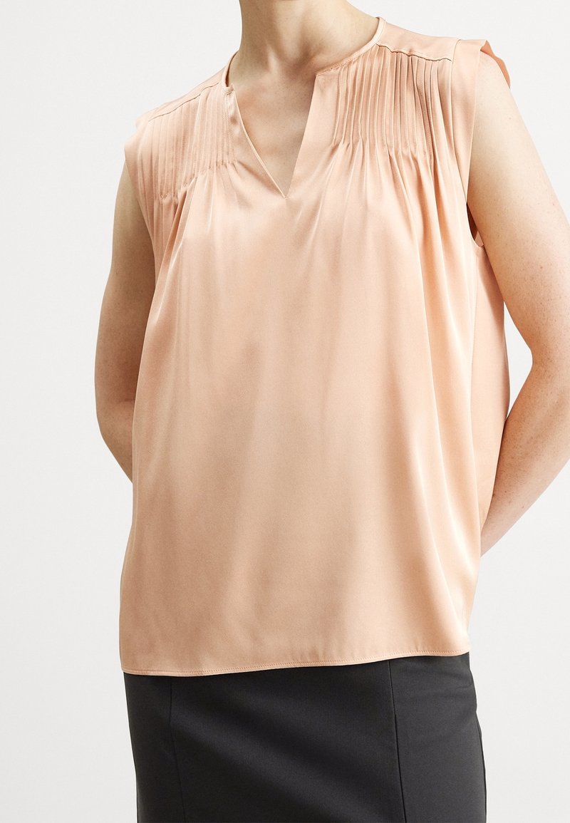 Blouse pêche sans manches avec détails plissés aux épaules et encolure en V, portée avec une jupe noire sur une personne debout.