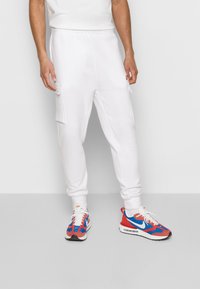 Vita joggers med sidofickor, smalnande med fåll vid anklarna, tillsammans med färgglada sneakers med röda, blå och vita detaljer.