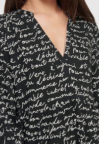 Vestido negro con escote en V, que presenta patrones de texto manuscrito en blanco. La tela parece ligera y fluye suavemente en el dobladillo.