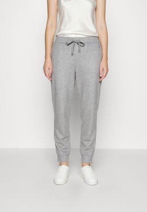 Persona vestida con pantalones tipo jogger gris claro con cordón, zapatillas blancas y una camiseta blanca sin mangas, de pie frente a un fondo liso y claro.