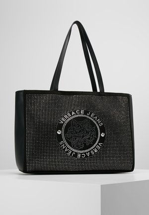 Czarna torba typu tote z srebrnymi ćwiekami i okrągłym logo Versace Jeans z dokładnym wizerunkiem głowy tygrysa na środku.