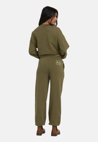 Kobieta z długimi ciemnymi włosami, ubrana w oliwkową bluzę i dopasowane dresy z logo na tylnej kieszeni, stoi z rękami na biodrach.