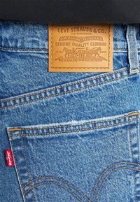Vaqueros de mezclilla azul con una etiqueta de cuero marrón que presenta el logo "LEVI STRAUSS & CO.", diseño remachado y una etiqueta roja de "Levi's" en el bolsillo trasero.