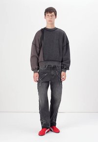 Sudadera negra de gran tamaño con mangas texturizadas y bordes deshilachados, combinada con jeans oscuros desgastados y zapatillas rojas. Detalle de capas sutiles.
