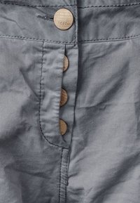 Un tissu gris au toucher lisse présente quatre grands boutons ronds en ton doré sur le devant, contrastant avec la couleur du matériau.
