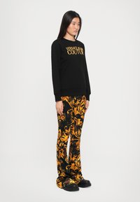 Čierna mikina so zlatým textom "VERSACE JEAN COUTURE". Skombinovaná s nohavicami s čiernym a žltým vzorom. Model má na sebe čierne robustné topánky.