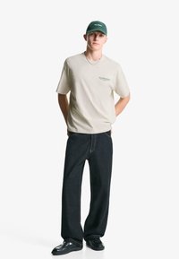 Tricou bej cu mânecă scurtă, cu text verde, asortat cu pantaloni largi din denim închis. Completat cu încălțăminte neagră și o șapcă verde.