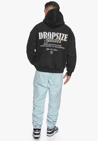 Black hoodie with a large white print on the back, paired with light blue jeans and black sneakers with white accents.