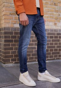 Jeans de denim azul ajustados con desgastes, combinados con zapatillas blancas con acentos texturizados. Llevado con una camisa naranja de botones.