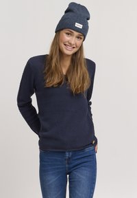 Pullover navy in tessuto testurizzato, maniche lunghe, scollatura rotonda e un piccolo logo decorativo. Abbinato a jeans blu e un berretto coordinato.