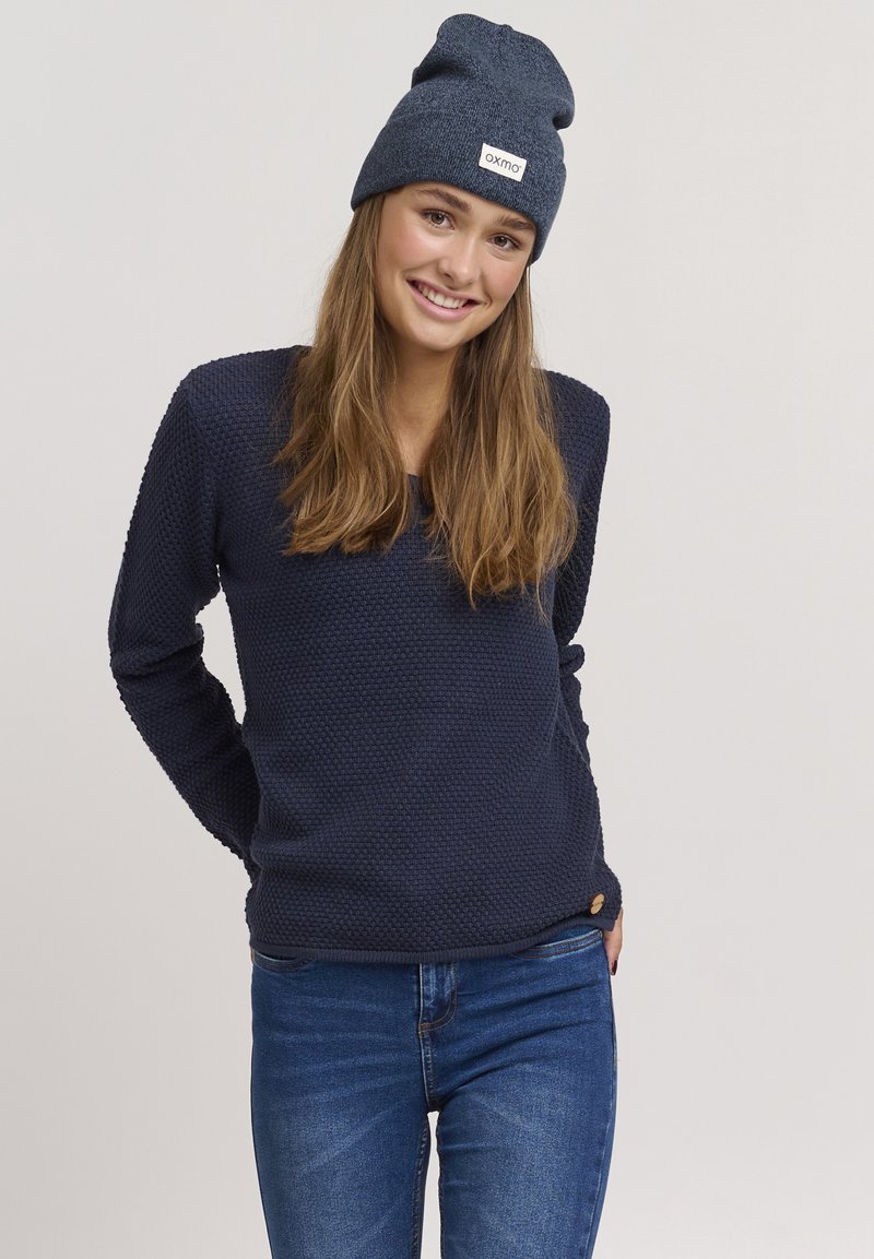 Pullover navy in tessuto testurizzato, maniche lunghe, scollatura rotonda e un piccolo logo decorativo. Abbinato a jeans blu e un berretto coordinato.