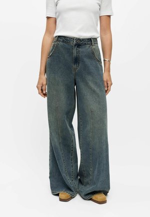 Personne portant un jean bleu délavé taille haute à jambes larges, une chemise blanche à manches courtes, des chaussures marron à bout carré et un bracelet argenté au poignet gauche.