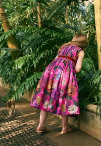 Fille en robe fleurie éclatante avec ceinture rouge se penche pour regarder des plantes dans un jardin intérieur luxuriant avec de grandes feuilles vertes.