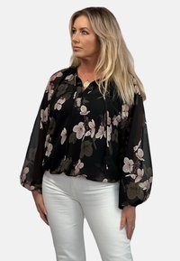 Czarna bluzka w kwiaty z długimi, zwiewnymi rękawami, z różowymi i zielonymi wzorami kwiatowymi, dekoltem w kształcie litery V oraz sznurkami do regulacji przy kołnierzyku.