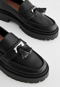 Black leather loafers with a textured finish, featuring a silver accent and tassels, and a thick black rubber sole.