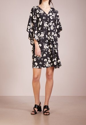 Kobieta ubrana w czarną sukienkę w kwiaty z dekoltem w serek, szerokimi rękawami oraz czarne sandały na słupku z odkrytymi palcami, stojąca na beżowej podłodze.