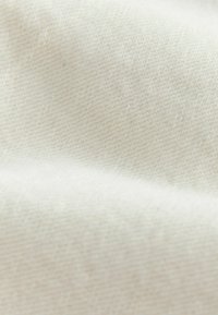 Közelkép puha, krémszínű anyagról, finoman szőtt textúrával és lágy redőkkel, amelyek sima felületet alkotnak.