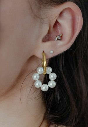 Goldene Creolen mit einem kreisförmigen Design, verziert mit weißen Perlen, ergänzt durch einen kleinen Perlenstecker und einem dekorativen Kristallohrring.