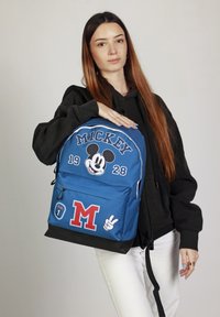 Mochila de tela azul con gráficos de Mickey Mouse, gran logotipo "M" y parches adicionales; incluye cierres de cremallera y una base negra.