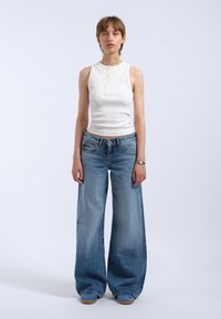 Top sans manches côtelé blanc associé à un jean en denim large bleu clair, à taille haute et légèrement délavé, maintenu en place par une ceinture.