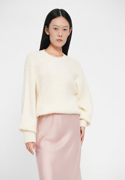 Kötött elefántcsont pulóver, amely kábelmintákkal és oversized ujjakkal rendelkezik, sima pasztell rózsaszín selyem szoknyával párosítva.